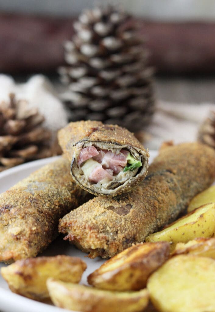 Three breaded, fried parasol mushroom rolls on a plate together with potato halves with two pine cones and rustic fabric in the background.